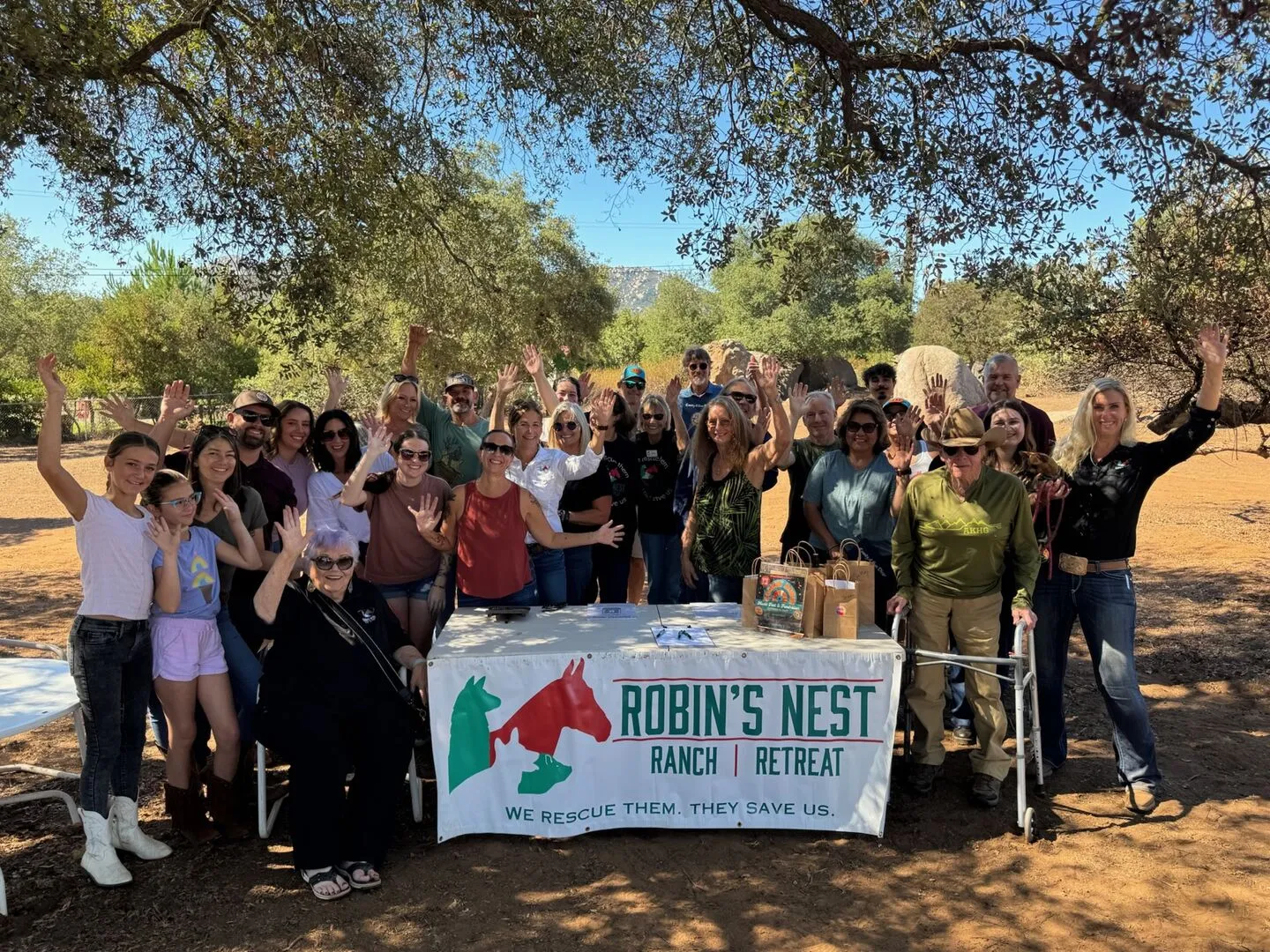 A large group of supporters smile and wave behind a Robin's Nest Ranch | Retreat banner under oak trees at a sanctuary gathering.